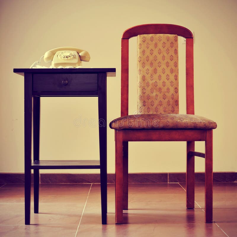 A beige old rotary dial telephone on a table next to a chair, with a retro effect. A beige old rotary dial telephone on a table next to a chair, with a retro effect