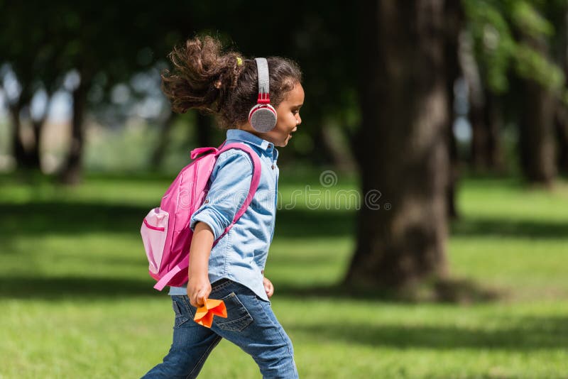 Adorable african american schoolkid in headphones wearing backpack and holding paper plane while walking in park. Adorable african american schoolkid in headphones wearing backpack and holding paper plane while walking in park