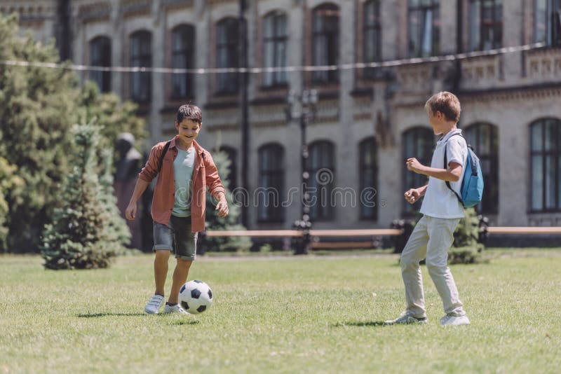 Two cheerful schoolboys with backpacks playing football on lawn near school. Two cheerful schoolboys with backpacks playing football on lawn near school