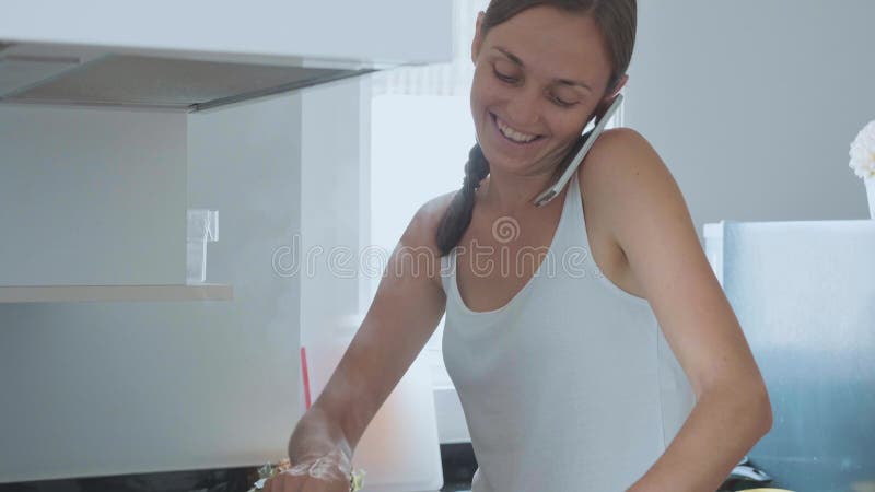 Ama de casa joven que cocina la cena, baile y hablando por el teléfono móvil