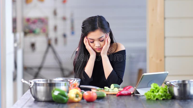 Ama de casa de sexo femenino asiática triste de moda que cocina la comida fresca en la cocina que tiene dolor de cabeza
