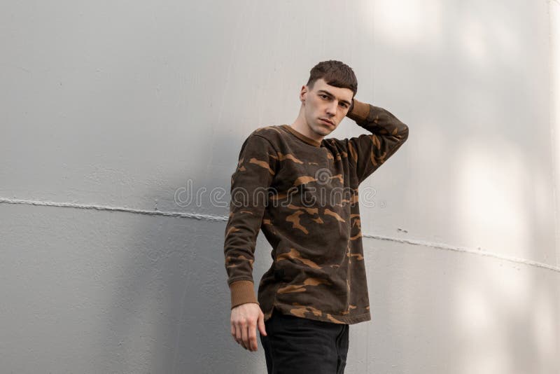 American handsome young man in fashionable military clothes with a stylish hairstyle poses near a metallic gray wall outdoors. stock photos American handsome young man in fashionable military clothes with a stylish hairstyle poses near a metallic gray wall outdoors. stock photos
