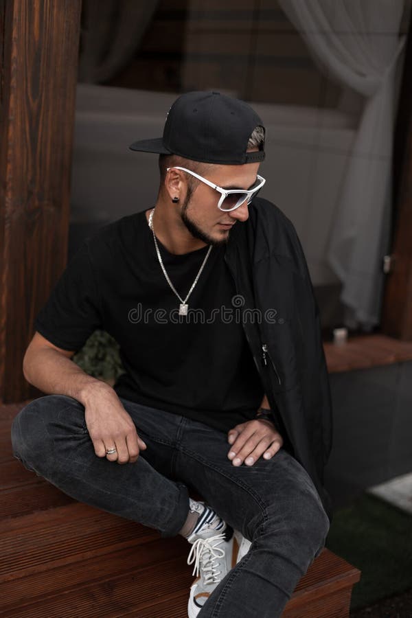 American hipster young man with a beard in a cap in a T-shirt in a jacket in sunglasses sits on wooden stairs near a vintage cafe royalty free stock photos American hipster young man with a beard in a cap in a T-shirt in a jacket in sunglasses sits on wooden stairs near a vintage cafe royalty free stock photos