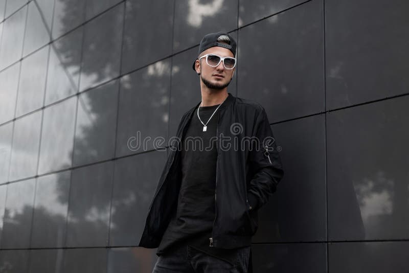 American young man with a beard in a cap in a t-shirt in a jacket in sunglasses is standing near a gray wall. Handsome hipster guy royalty free stock photos American young man with a beard in a cap in a t-shirt in a jacket in sunglasses is standing near a gray wall. Handsome hipster guy royalty free stock photos