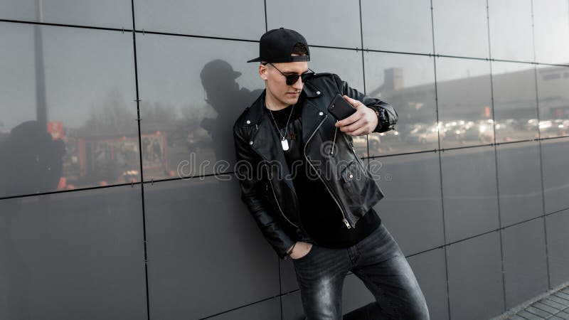 American young man hipster in a black leather jacket in a baseball cap in jeans in a t-shirt is standing near the modern building stock photo American young man hipster in a black leather jacket in a baseball cap in jeans in a t-shirt is standing near the modern building stock photo