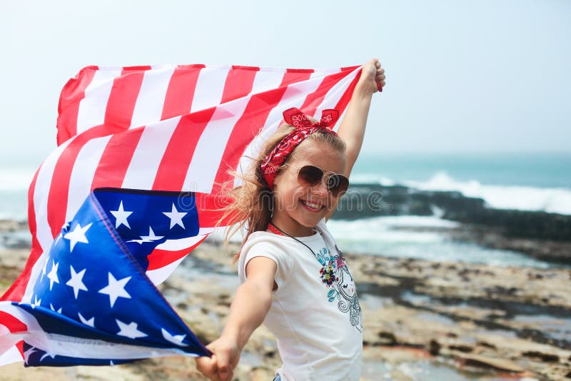 American flag. Little smiling patriotic girl with long blond hair, red head band bandana and sunglasses holding an american flag waving in the wind on the ocean beach. National 4 july. Memorial day. American flag. Little smiling patriotic girl with long blond hair, red head band bandana and sunglasses holding an american flag waving in the wind on the ocean beach. National 4 july. Memorial day