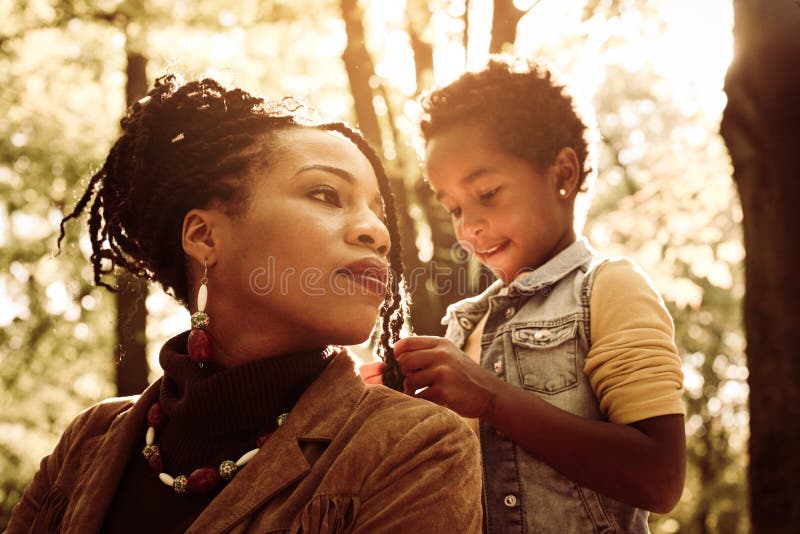 Daughter making mom`s hairstyle in the park. African American women with daughter in park. Daughter making mom`s hairstyle in the park. African American women with daughter in park.