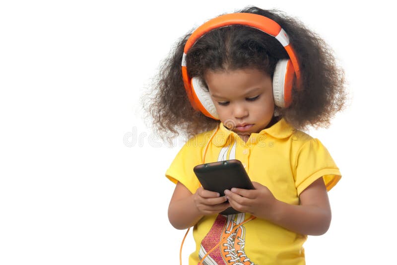 Cute african american small girl listening to music on a cellphone using big orange headphones isolated on white. Cute african american small girl listening to music on a cellphone using big orange headphones isolated on white