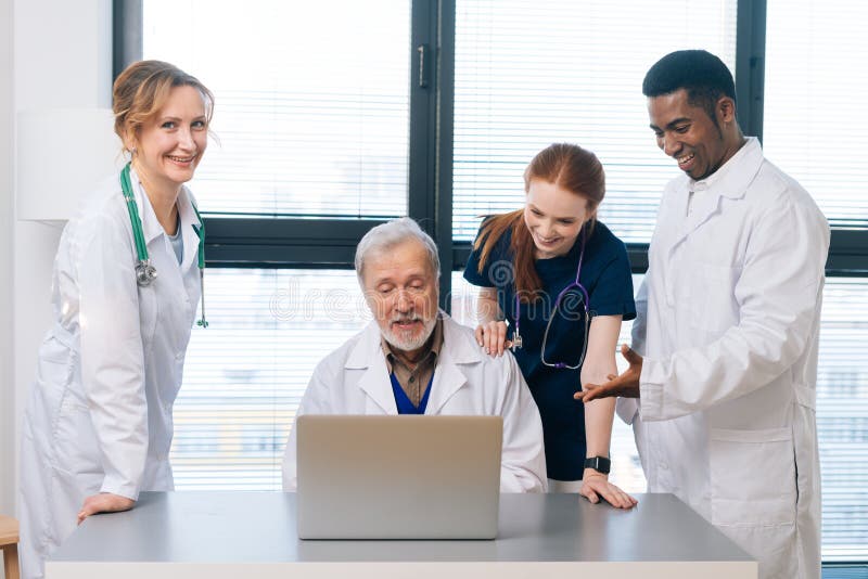 Amichevole team multietnico di medici uomini e donne a tavola con briefing sorridendo guardando lo schermo del portatile e chiacchierando sullo sfondo della finestra. gruppo di medici che riposano in ospedale.