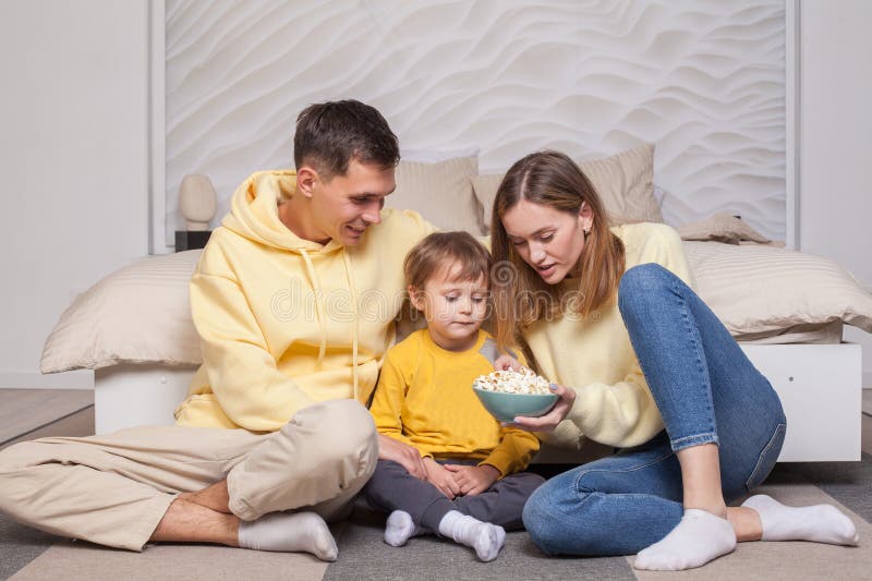Amichevoli genitori con figli figlio seduti vicino al letto e tengono in mano una ciotola con dei popcorn fotografia stock