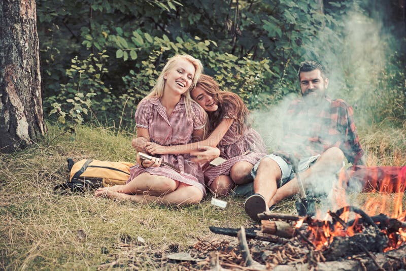 Amici allegri che hanno picnic in legno Uomo barbuto e due libri di lettura sorridenti delle ragazze da fuoco di accampamento, so fotografia stock libera da diritti Amici allegri che hanno picnic in legno Uomo barbuto e due libri di lettura sorridenti delle ragazze da fuoco di accampamento, so fotografia stock libera da diritti