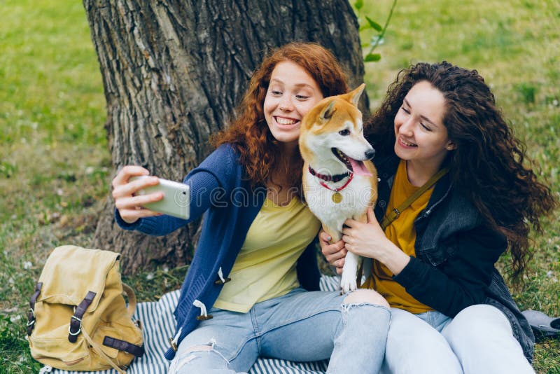 Amici di ragazze felici che prendono selfie in parco con il cane sveglio facendo uso della macchina fotografica dello smartphone immagini stock libere da diritti Amici di ragazze felici che prendono selfie in parco con il cane sveglio facendo uso della macchina fotografica dello smartphone immagini stock libere da diritti