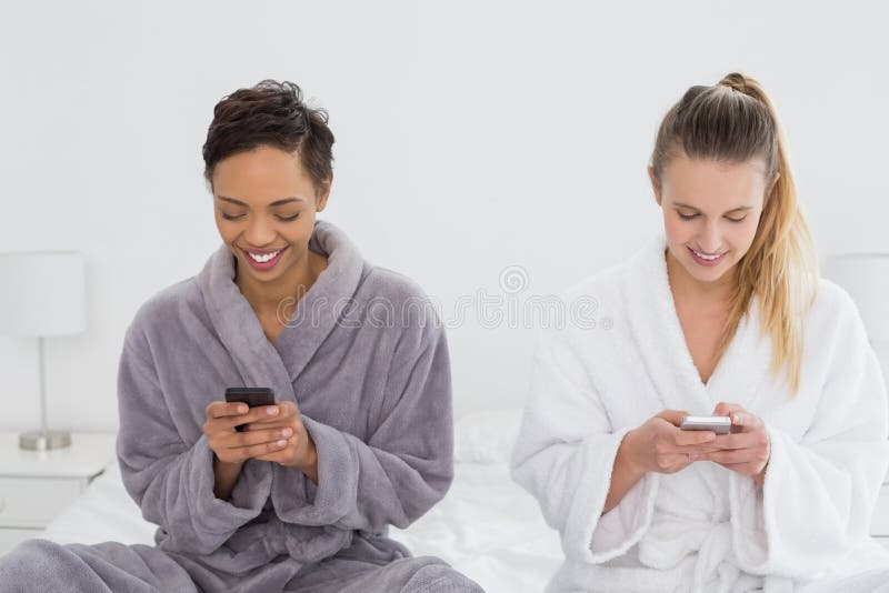 Amici femminili nell'invio di messaggi di testo degli accappatoi sul letto immagine stock libera da diritti Amici femminili nell'invio di messaggi di testo degli accappatoi sul letto immagine stock libera da diritti