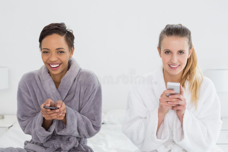 Amici femminili nell'invio di messaggi di testo degli accappatoi sul letto fotografie stock