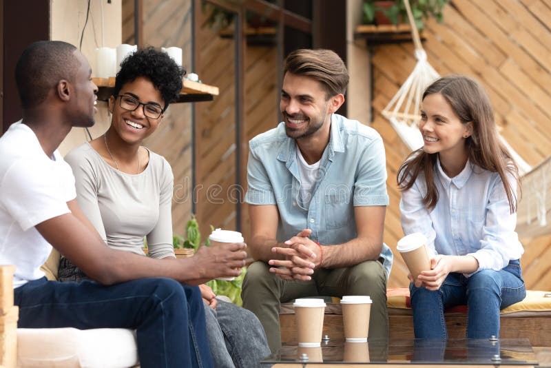 Four laughing diverse people positive multi-ethnic friends sitting in cozy cafe summer terrace drink coffee telling funny stories from life feels happy and satisfied enjoy time together on weekend. Four laughing diverse people positive multi-ethnic friends sitting in cozy cafe summer terrace drink coffee telling funny stories from life feels happy and satisfied enjoy time together on weekend.