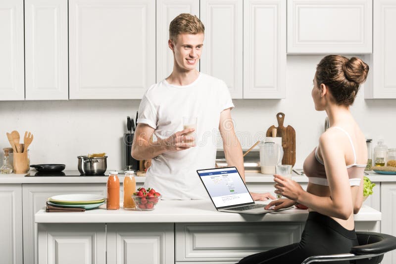 girlfriend using laptop with loaded facebook page and looking at boyfriend in kitchen. girlfriend using laptop with loaded facebook page and looking at boyfriend in kitchen