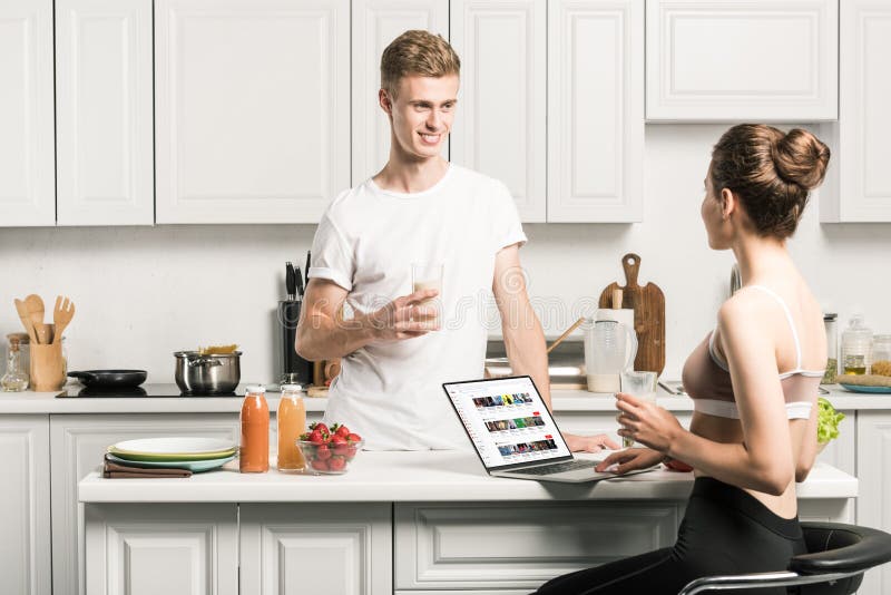 girlfriend using laptop with loaded youtube page and looking at boyfriend in kitchen. girlfriend using laptop with loaded youtube page and looking at boyfriend in kitchen