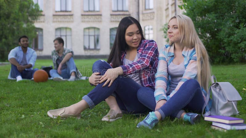 Amigos bonitos da misturado-raça que sentam-se no gramado e que discutem vestidos novos, estudantes