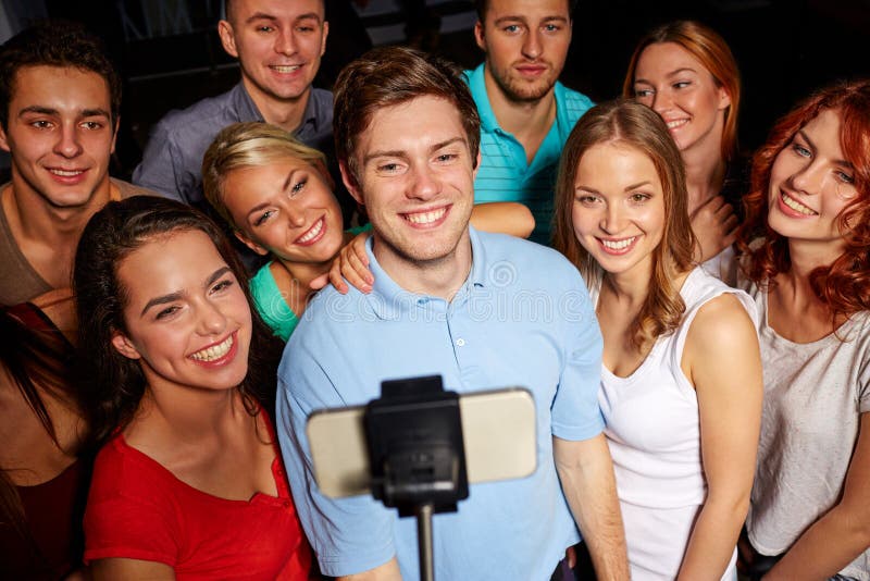 Party, technology, nightlife and people concept - smiling friends with smartphone and monopod taking selfie in club. Party, technology, nightlife and people concept - smiling friends with smartphone and monopod taking selfie in club