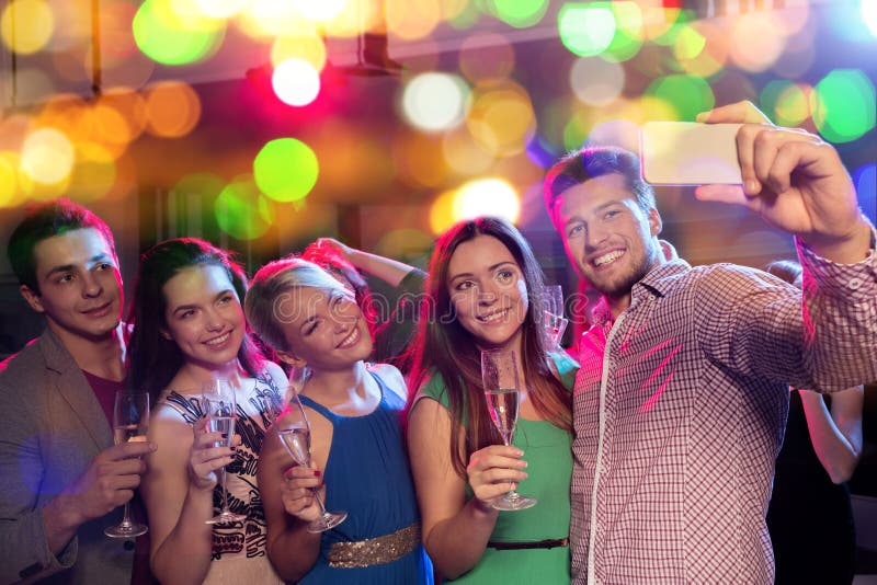 Party, holidays, technology, nightlife and people concept - smiling friends with glasses of non-alcoholic champagne and smartphone taking selfie in club. Party, holidays, technology, nightlife and people concept - smiling friends with glasses of non-alcoholic champagne and smartphone taking selfie in club