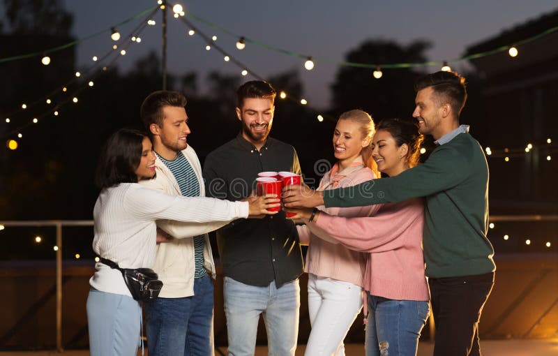 Amigos que tintinean las tazas del partido en tejado en la noche imagenes de archivo Amigos que tintinean las tazas del partido en tejado en la noche imagenes de archivo