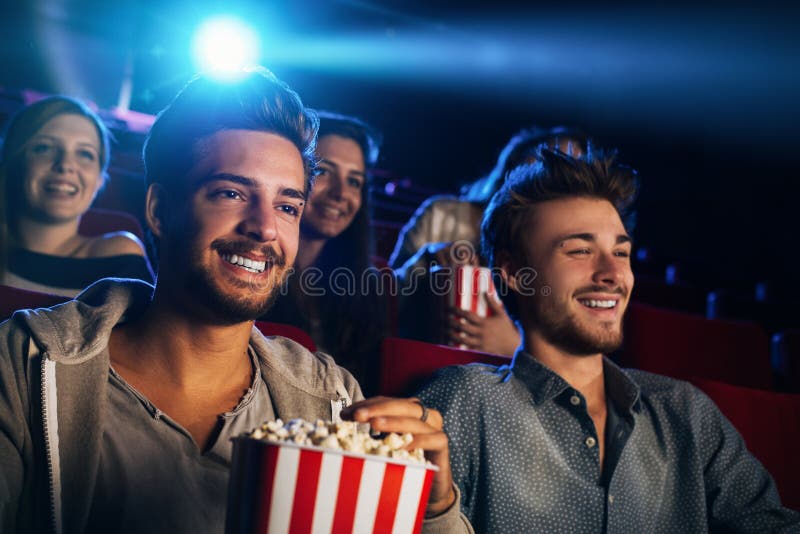 Young people sitting at the cinema, watching a movie and eating popcorn, two smiling men on foreground. Young people sitting at the cinema, watching a movie and eating popcorn, two smiling men on foreground