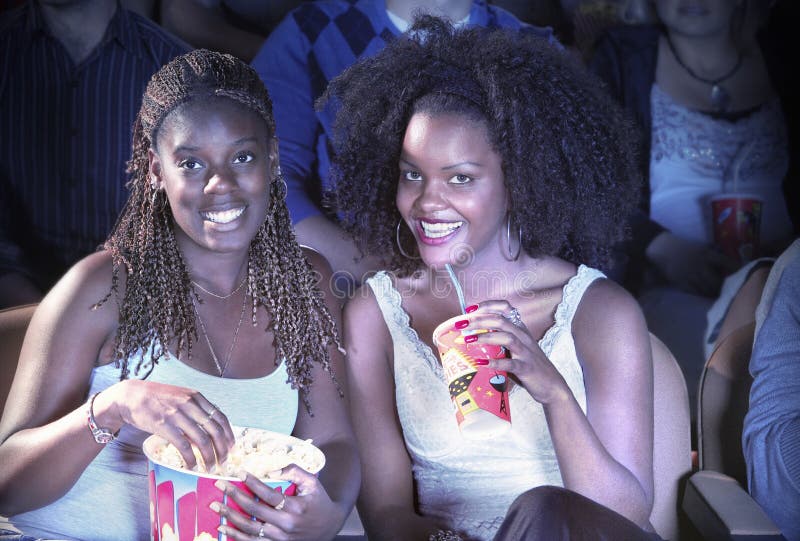 Portrait of smiling African American female friends with drink and popcorn watching movie in theater. Portrait of smiling African American female friends with drink and popcorn watching movie in theater