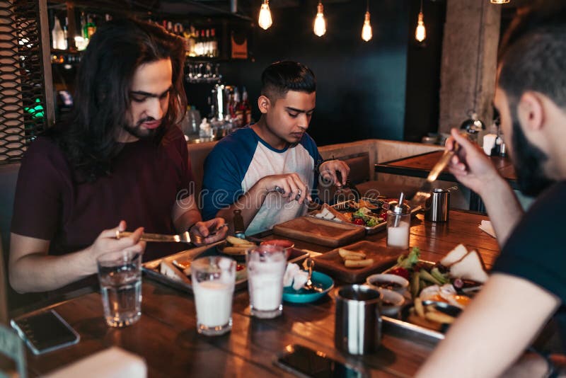 Amis multiraciaux mangeant le petit déjeuner en café Les jeunes hommes causent tout en ayant la nourriture et les boissons savour photographie stock Amis multiraciaux mangeant le petit déjeuner en café Les jeunes hommes causent tout en ayant la nourriture et les boissons savour photographie stock