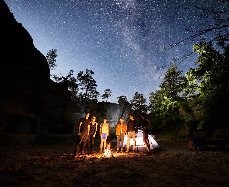 Friends travellers standing together near bonfire beside camp and glowing tourist tent at night. On background beautiful night starry sky full of stars and Milky way, mountain rocks and trees. Friends travellers standing together near bonfire beside camp and glowing tourist tent at night. On background beautiful night starry sky full of stars and Milky way, mountain rocks and trees