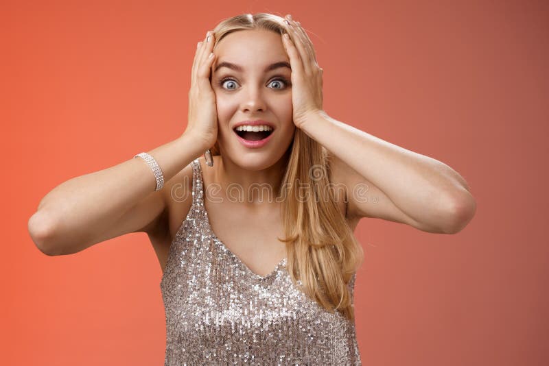 Amused surprised excited young blond attractive woman in silver glittering stylish dress hold head hands widen eyes