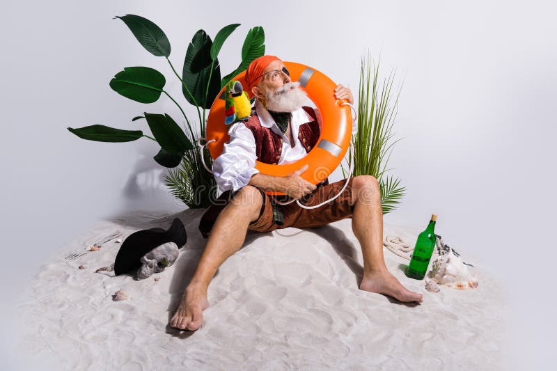 Amusing scene of a bearded pirate posing on sandy beach with parrot and lifebuoy, surrounded by tropical foliage and stock photo