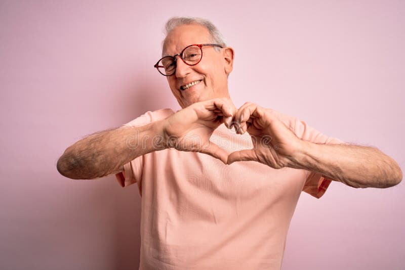 Anciano de pelo gris con gafas parado sobre fondo rosado aislado sonriendo enamorado mostrando símbolo del corazón y forma con