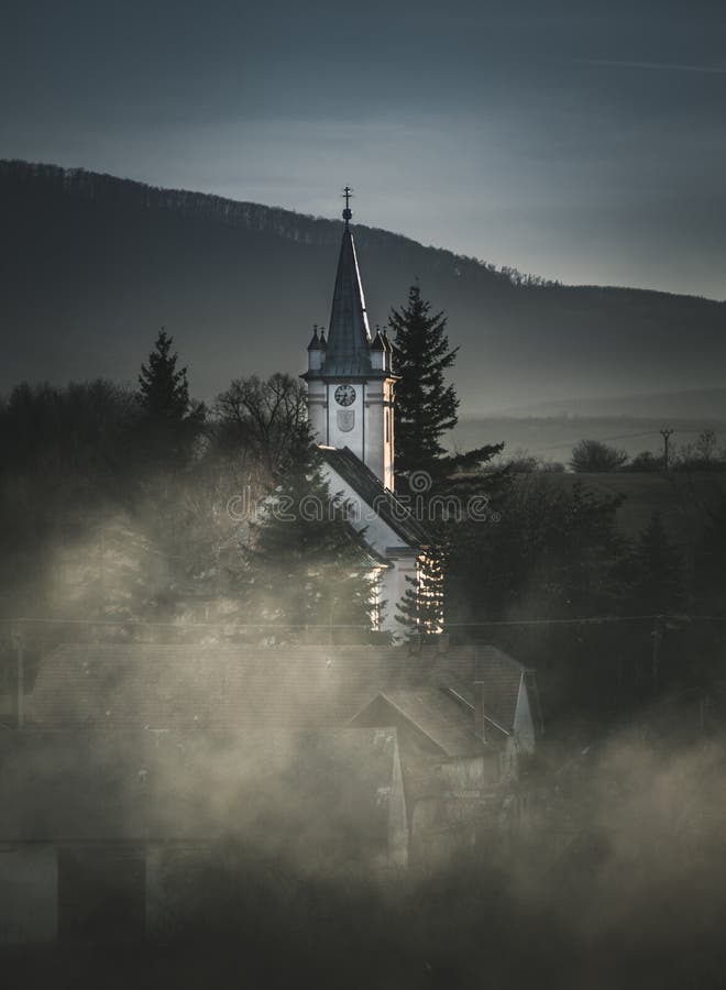 Ancient church in village shrouded in fog with big mountains and hills on background. Photo of White church from the green meadow