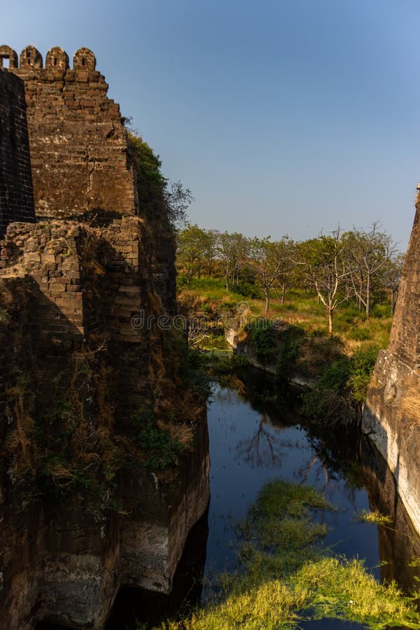 Ancient stone fortifications rise above a deep, reflective water-filled moat. The walls are weathered and partially covered with vegetation. Beyond the moat, dense greenery and sparse trees create a lush backdrop under a clear blue sky. The fort's strategic construction is evident in the use of natural and man-made defenses. The scene conveys a sense of historical significance and architectural ingenuity, typical of structures designed for defense and protection. Sparse use stock images, royalty-free photos and pictures