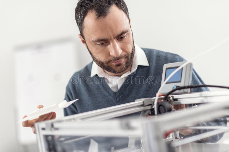 Necessary checkup. Charming bearded young man holding a recently printed 3D model and looking inside the 3D printer, checking its work. Necessary checkup. Charming bearded young man holding a recently printed 3D model and looking inside the 3D printer, checking its work