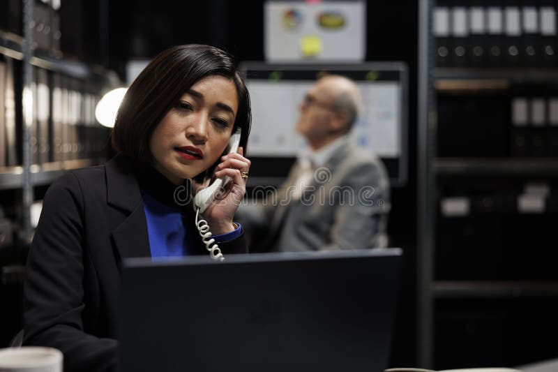 Bookkeeping businesswoman offering consultancy during phone call in administrative workplace. Asian employee in archival depository filled with document folders and flowcharts. Bookkeeping businesswoman offering consultancy during phone call in administrative workplace. Asian employee in archival depository filled with document folders and flowcharts