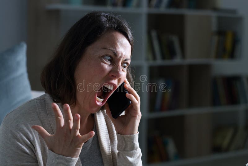 Angry adult woman screaming calling on smart phone sitting in the livingroom at night at home. Angry adult woman screaming calling on smart phone sitting in the livingroom at night at home
