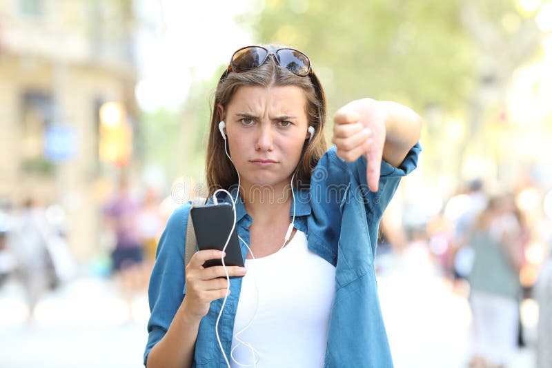 Front view portrait of an annoyed girl listening to music with thumbs down in the street