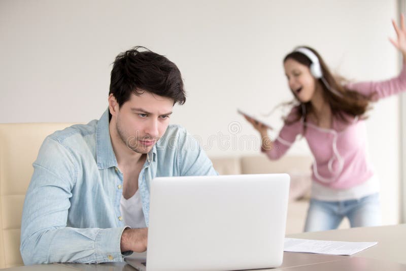 Busy guy working on laptop computer, annoyed by girlfriend listening to music on tablet, dancing near him. Working at home problems, too noisy to study, men can t work because of bothering loud girl. Busy guy working on laptop computer, annoyed by girlfriend listening to music on tablet, dancing near him. Working at home problems, too noisy to study, men can t work because of bothering loud girl