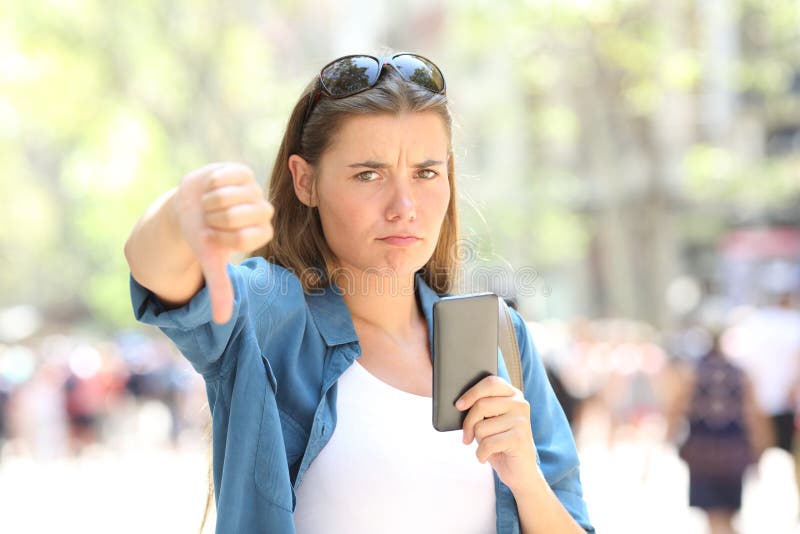 Annoyed woman holding a smart phone looking at camera with thumbs down in the street. Annoyed woman holding a smart phone looking at camera with thumbs down in the street