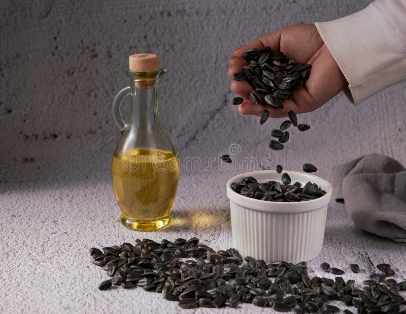 An anonymous woman`s hand pours sunflower seeds into a white ceramic bowl and loose sunflower seeds on a table with a glass bottl royalty free stock photography