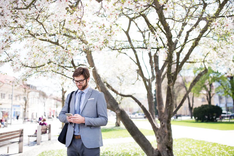 Handsome businessman scrolling on smartphone while standing on city street, going to office. Manager smiling, outdoor in urban setting. Handsome businessman scrolling on smartphone while standing on city street, going to office. Manager smiling, outdoor in urban setting.