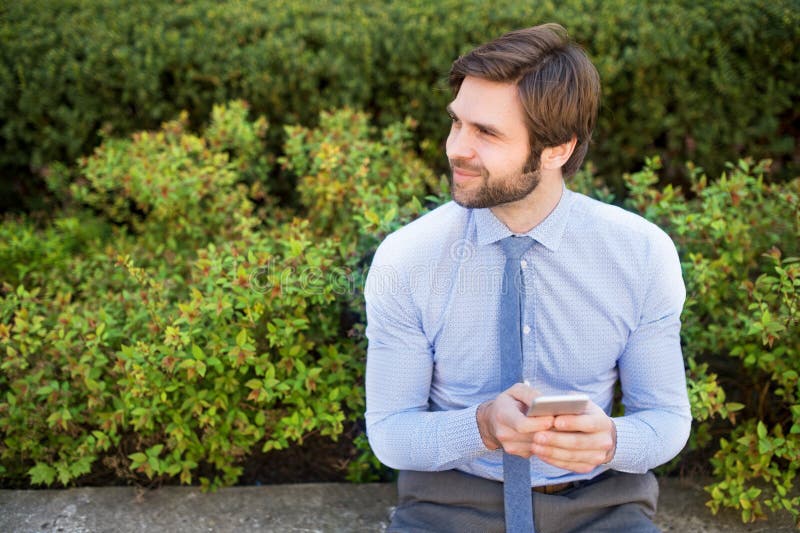 Handsome businessman holding smartphone sitting on bench in the city park. Working remotely, waiting for business meeting. Manager smiling, outdoor in urban setting. Handsome businessman holding smartphone sitting on bench in the city park. Working remotely, waiting for business meeting. Manager smiling, outdoor in urban setting