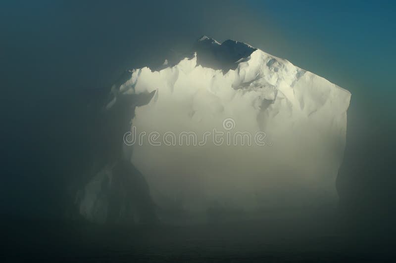 A large frontlit Antarctic iceberg emerges from the morning mists through which a blue sky is shimmering in the background. A large frontlit Antarctic iceberg emerges from the morning mists through which a blue sky is shimmering in the background.