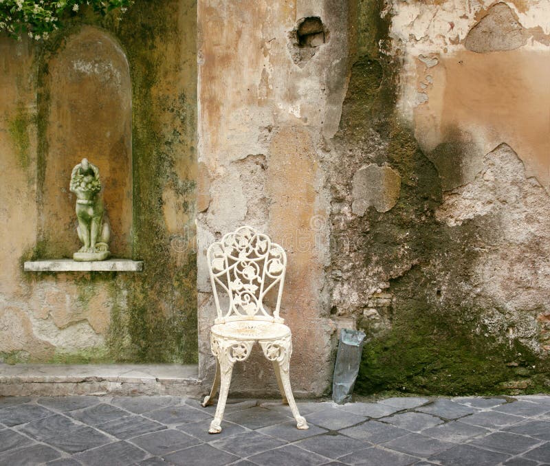 A characteristic and decadent corner of an old crumbling wall with an old wrought iron chair and a little statue. A characteristic and decadent corner of an old crumbling wall with an old wrought iron chair and a little statue
