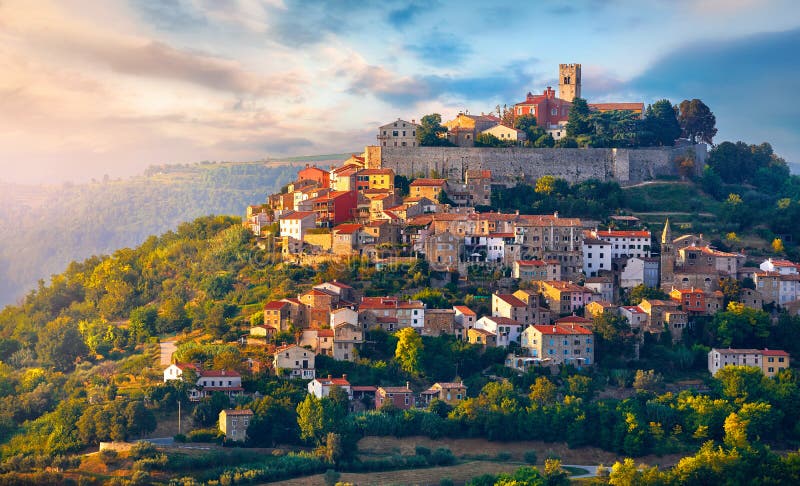 Antique city Motovun Croatia Istria. Picturesque panorama age-old village at hill with pink cloud and sunny light and authentic home with red tegular roof and green vineyard garden. Antique city Motovun Croatia Istria. Picturesque panorama age-old village at hill with pink cloud and sunny light and authentic home with red tegular roof and green vineyard garden.