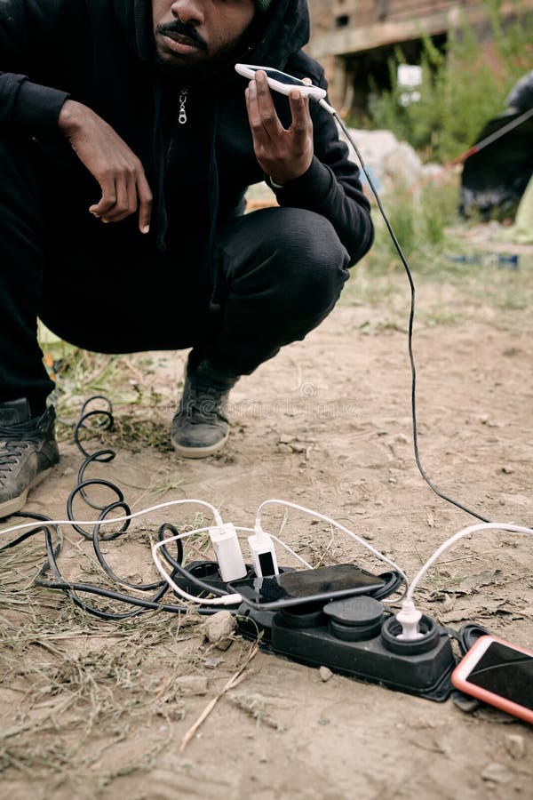 Close-up of black refugee man crouching at socket outlet and listening to audio message on phone while charging it outdoors. Close-up of black refugee man crouching at socket outlet and listening to audio message on phone while charging it outdoors