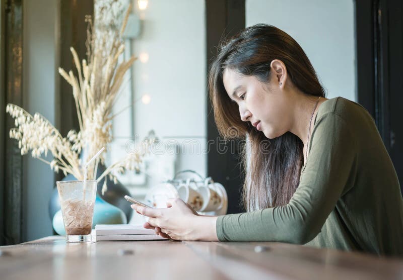 Använder den asiatiska kvinnan för closeupen en smart telefon och att dricka på med is choklad på träräknareskrivbordet i coffee arkivfoton Använder den asiatiska kvinnan för closeupen en smart telefon och att dricka på med is choklad på träräknareskrivbordet i coffee arkivfoton