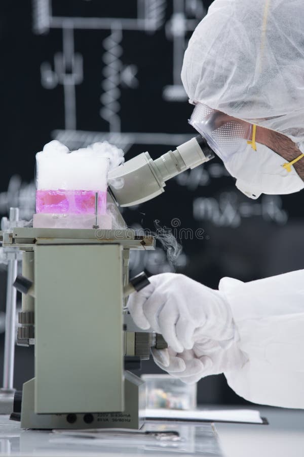 Close-up of scientist in a chemistry lab analyzing under microscope around lab tools and colorful liquids with a blackboard on the background. Close-up of scientist in a chemistry lab analyzing under microscope around lab tools and colorful liquids with a blackboard on the background