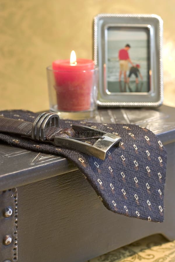 Man's belt and tie hang over a trinket box on this dresser vignette. Also visible in the background are a lit red candle and a blurred photo of a man and small boy at the beach. Gold paisley print surrounds the objects. Man's belt and tie hang over a trinket box on this dresser vignette. Also visible in the background are a lit red candle and a blurred photo of a man and small boy at the beach. Gold paisley print surrounds the objects.